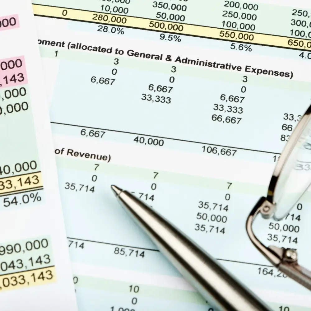 A close-up view of a hand pointing with a pen at numbers on a financial spreadsheet or chart, showing data for Revenue and General & Administrative expenses.