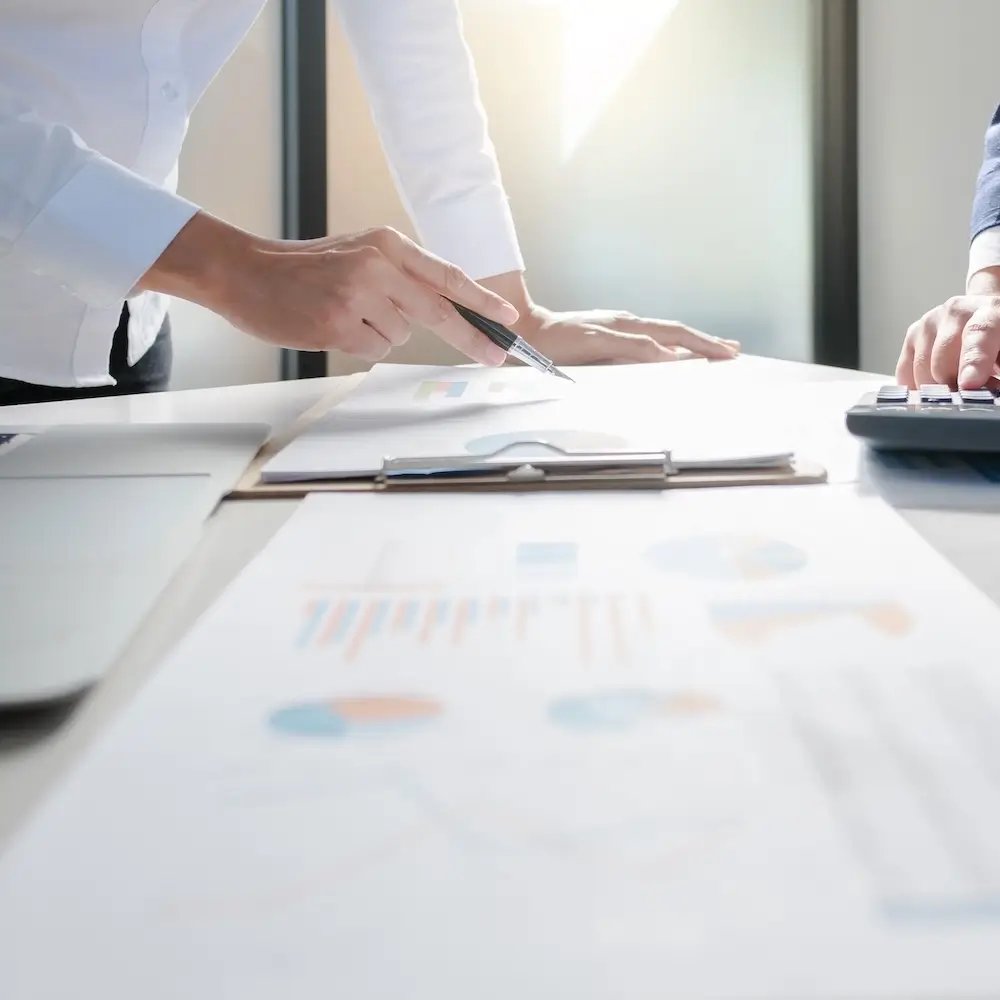A close-up view of two business colleagues meeting at a table, one person's hand is pointing at a document or chart with a pen, reviewing performance reports.