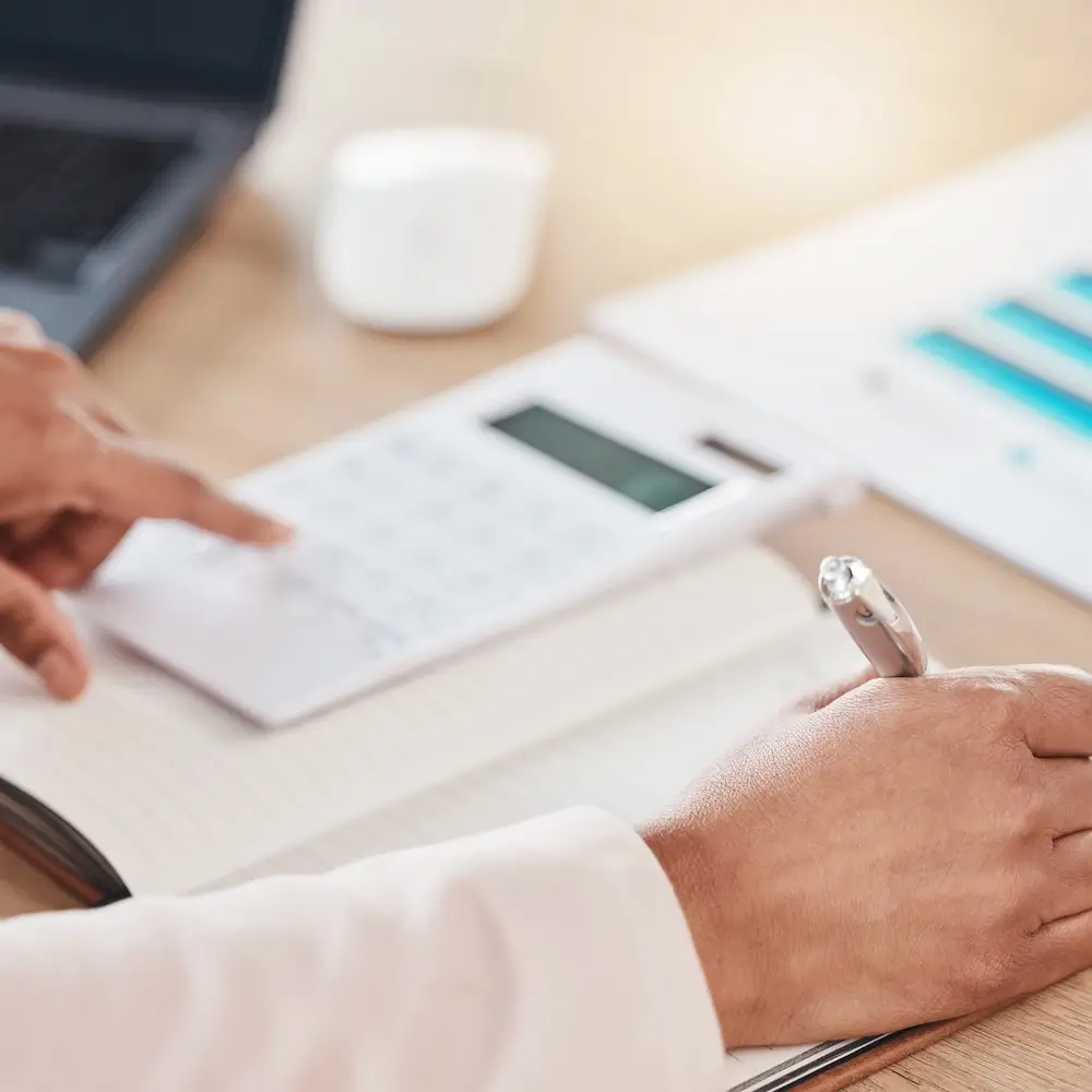 Close-up of a person's hands writing with a pen in the foreground while simultaneously using a calculator on a desk in the background, suggesting accounting or financial work.