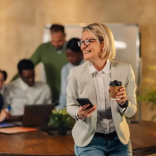 A smiling mature businesswoman standing in an office, holding a coffee cup and looking at her smartphone during a meeting with blurred colleagues in the background.