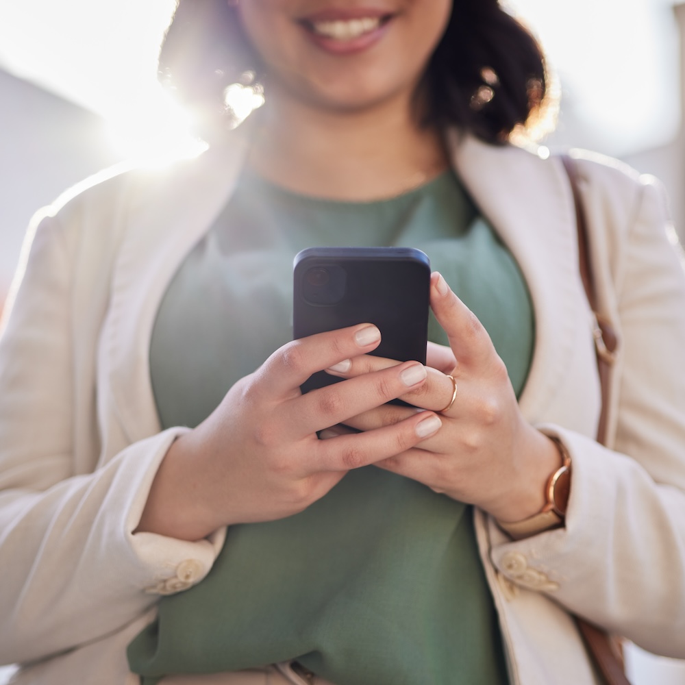 Close-up of a smiling woman's hands holding a black smartphone outdoors in a bright city setting, wearing a light jacket and a watch