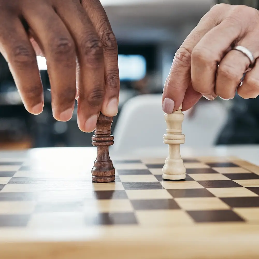 Close-up of two people's hands, one dark-skinned and one light-skinned, moving a dark king piece and a light queen piece on a wooden chessboard, symbolizing strategy or a crucial decision.