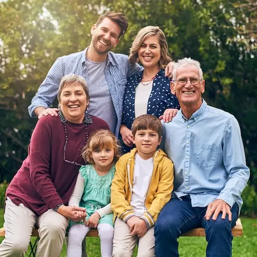 A smiling, multi-generational family of six sitting close together on an outdoor bench, including two grandparents, two parents, a young girl, and a young boy.