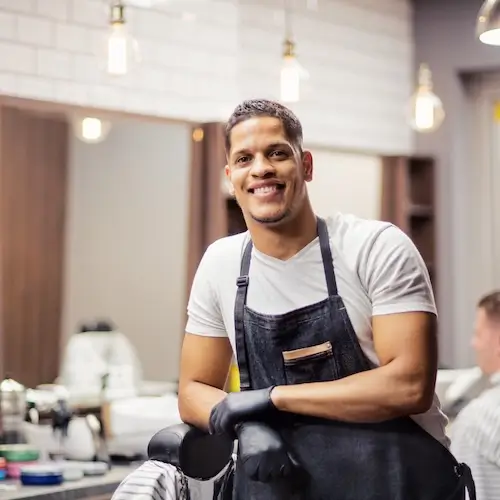 A smiling young Hispanic male barber or hairdresser standing with arms crossed in a salon, wearing a dark apron and black gloves, with salon equipment in the blurred background.