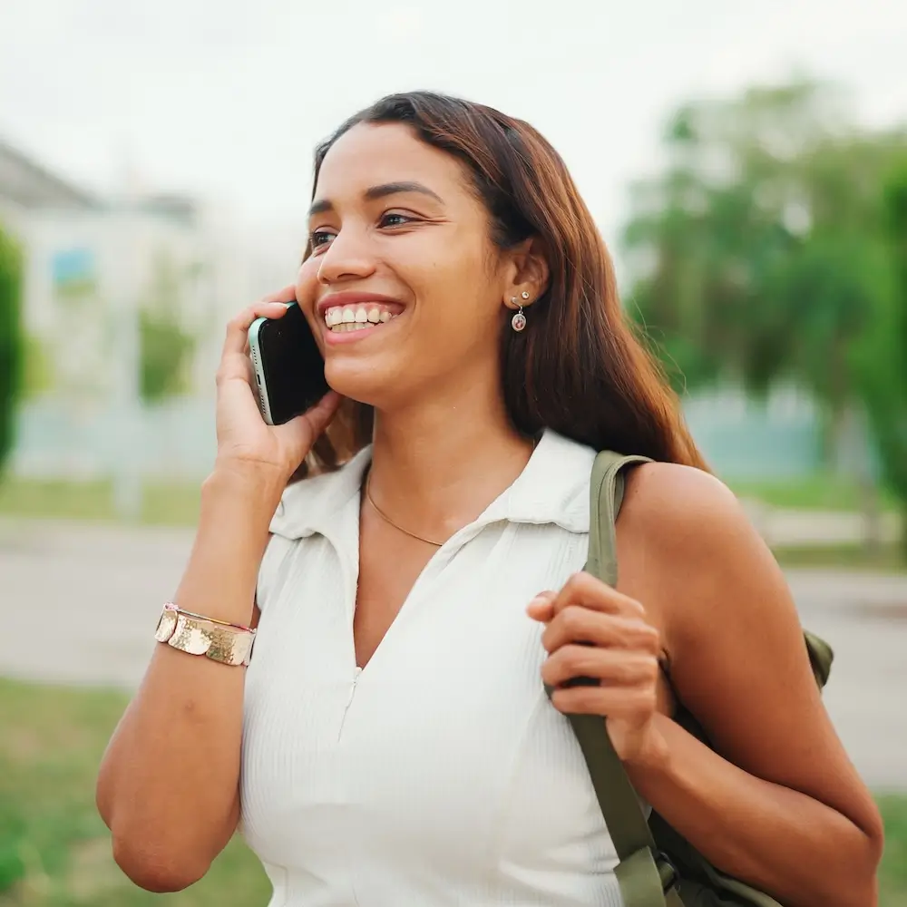 A young woman with brown hair, smiling brightly and talking on a black smartphone outdoors, wearing a light collared shirt and a gold bracelet.